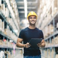 Smart Indian engineer man wearing safety helmet doing stock tick check and cardboard stock product management in factory warehouse background